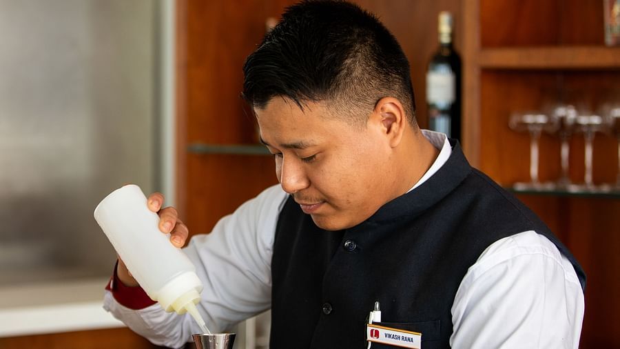 A bartender in uniform prepares a drink behind the counter, pouring liquid into a jigger, with bar tools and glasses neatly arranged at Mandala Norling Lords Prime in Kathmandu.