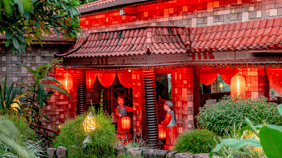 An exterior view of Ming Dynasty at Noor-Us-Sabah Palace, Bhopal, showing the traditional red-roofed building and glowing lanterns set behind a manicured green lawn.