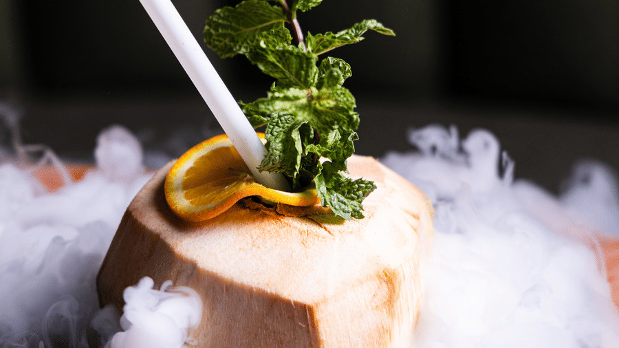 A tropical cocktail creatively presented in a coconut shell, topped with fresh mint and surrounded by smoky mist for an exotic and dramatic visual effect at Papaya Tree Hotels.