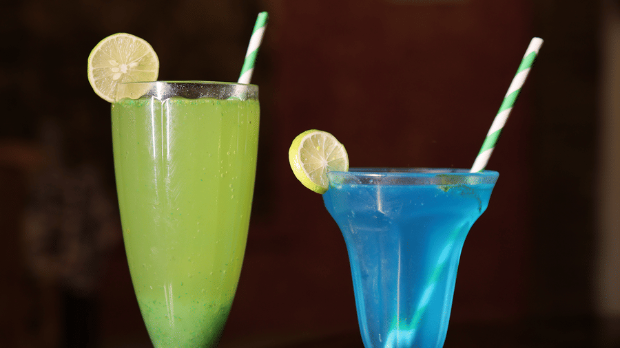 Two vibrant, layered mocktails in shades of green and blue, garnished with lemon slices and straws.