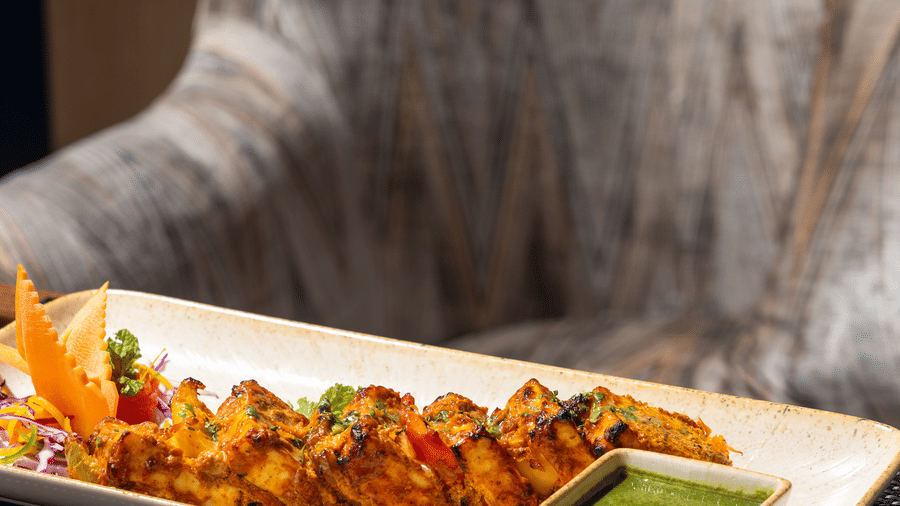 A rectangular ceramic platter of tandoori paneer tikka at Sands Point Hotel, served with a side of vibrant green mint chutney and a decorative carved carrot garnish, placed on a textured dark table runner against a striped upholstered chair.