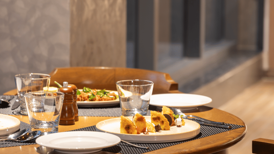 A neatly set round wooden dining table at Sands Point Hotel with water glasses, a pepper grinder, white side plates, and a plated dessert with golden fried pieces, surrounded by wooden chairs with upholstered seats.