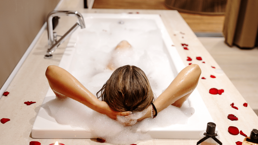 A person reclining in a rectangular bathtub with their arms behind their head at SUKAVI, surrounded by flower petals arranged around the bath area.