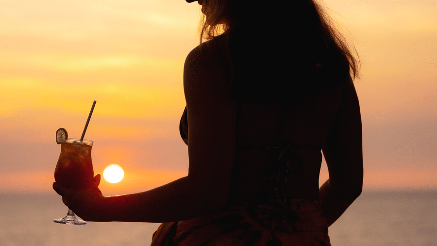 Silhouette of a person holding a drink with a straw beside the infinity pool at SUKAVI, with the ocean and sunset creating a scenic backdrop.
