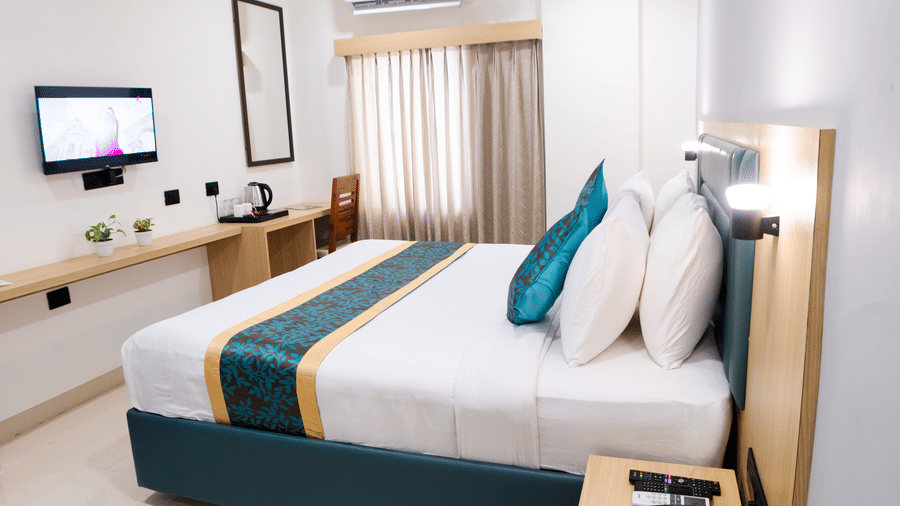 Side view of the room with a neatly made bed, bedside table with telephone and remotes, wall-mounted TV, and curtains covering the windows at Sunrise Business Hotel, Hyderabad.