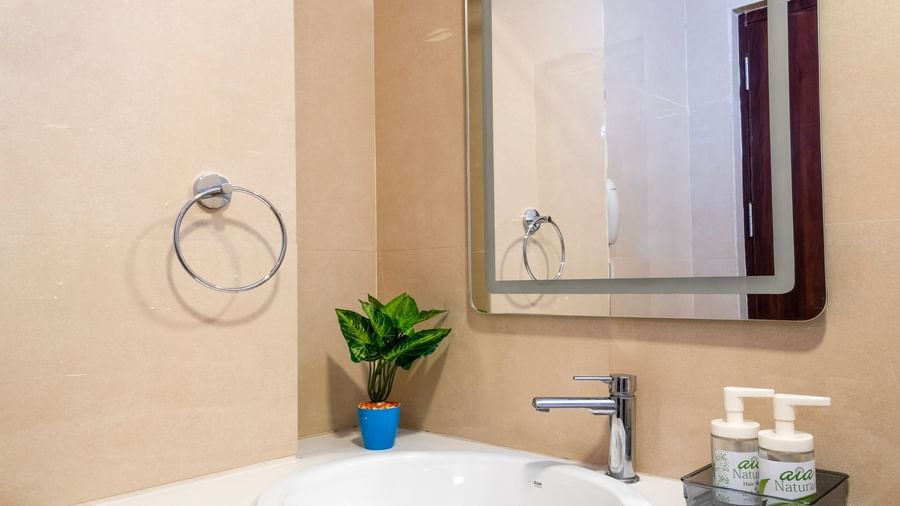 Close up of a bathroom wash basin with mirror and wall fixtures surrounded by beige tiles and a touch of greenery at Swarna's The Capital.