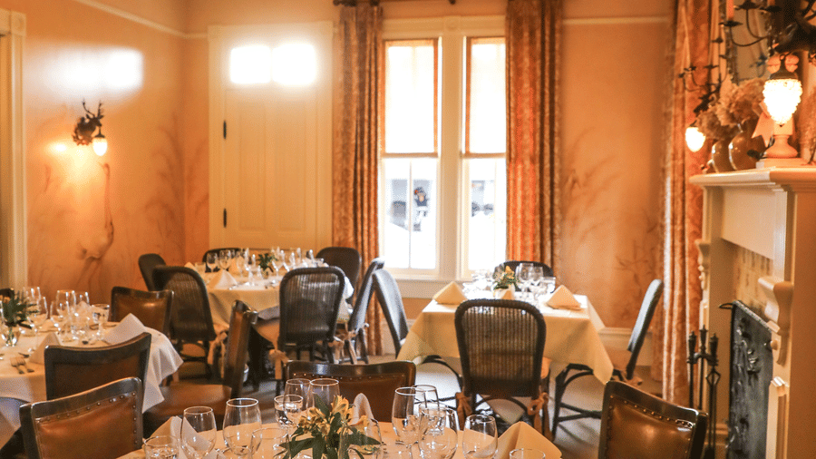 A round dining table with neatly folded napkins and wine glasses at Tallman Hotel, illuminated by a warm chandelier and surrounded by wooden chairs.