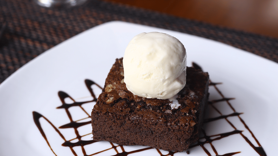 A vertical shot of a square chocolate brownie topped with a scoop of vanilla ice cream and a drizzle of chocolate sauce at The Bull Boutique Hotel.