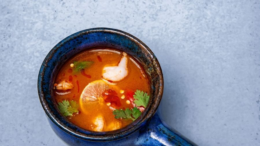 A bowl of soup with vegetables and herbs, served in a dark blue ladle-like bowl on a light textured surface at The Hamuse Hotel, Kodaikanal.