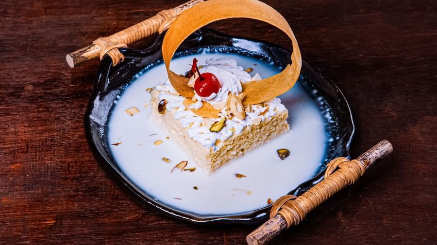 A plated dessert with a decorative basket element, garnished with cream and a cherry, presented on a white plate with sauce swirls on a dark wooden surface at The Hamuse Hotel, Kodaikanal.