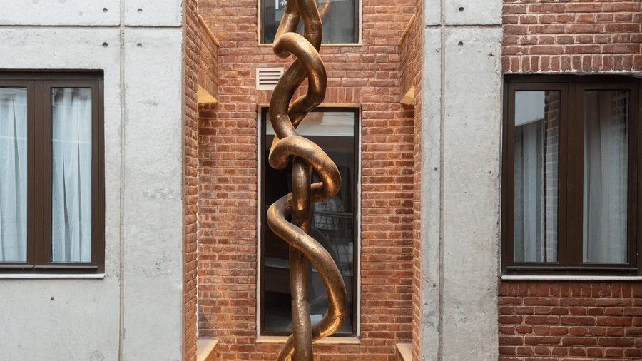 A tall, vertical view of an abstract metallic sculpture hanging between contemporary concrete and classic brick hotel walls in Nepal.