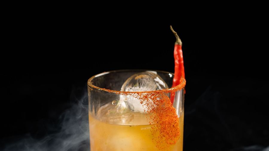 A cocktail glass filled with an amber-colored drink and an orange peel garnish is set against a dark background with smoke effects at The Qualia Resort Club & Brewery, Udaipur.