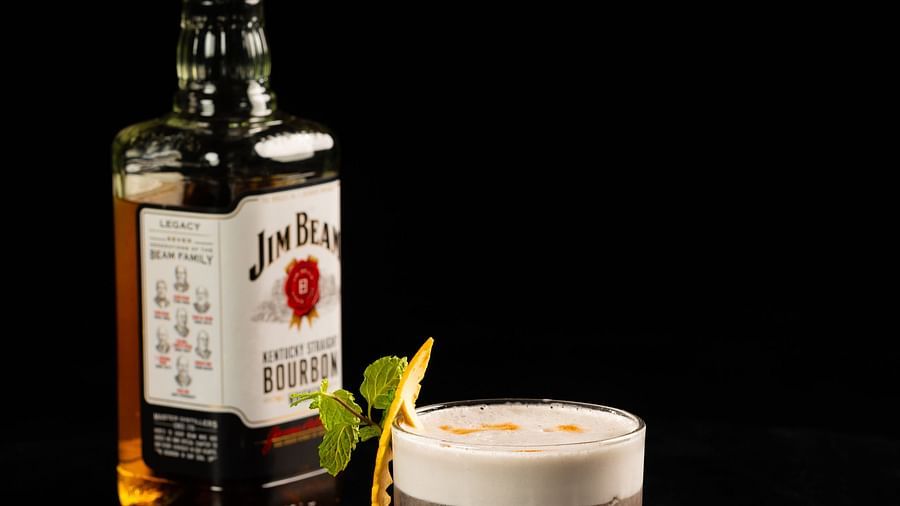 A bottle of syrup and a cocktail glass filled with a creamy beverage, garnished with a lime wedge, are placed on a sleek black surface at The Qualia Resort Club & Brewery, Udaipur.
