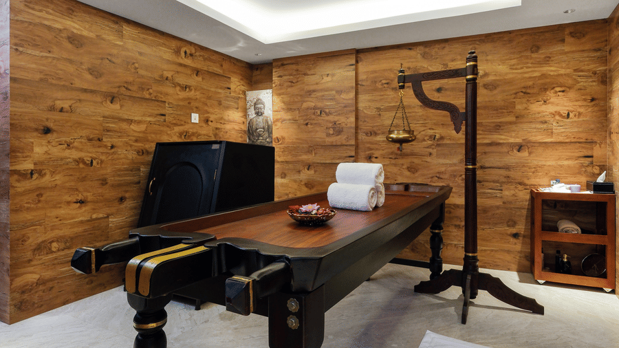 A traditional wooden Ayurveda massage table with a headrest stands in a wood-panelled wellness room beneath an air conditioning unit at Tiara by MPS.