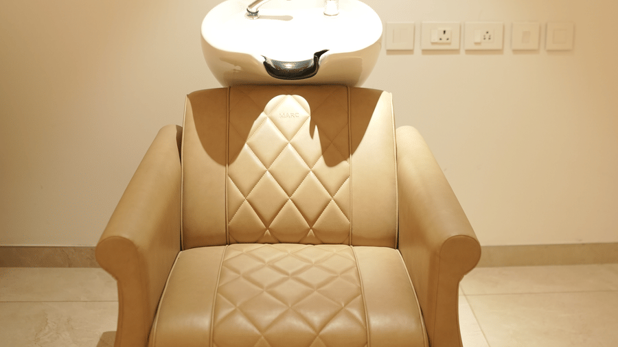 A beige leather hair washing chair with a white ceramic basin attached stands against a plain wall beneath a framed picture at Tiara by MPS.