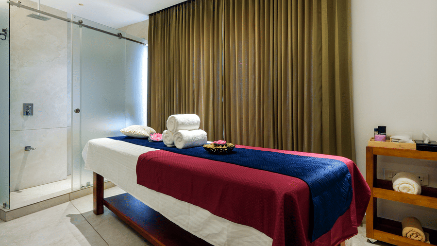 An elegant spa treatment room featuring a central massage table with folded towels, a storage cart, and an adjacent glass walled shower at Tiara by MPS.