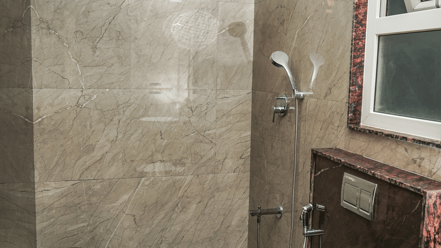 Modern bathroom featuring neutral stone-effect tiles, a wall-mounted toilet, a chrome shower head, and a white ceramic basin at Trident The Boutique Hotel, Ooty.