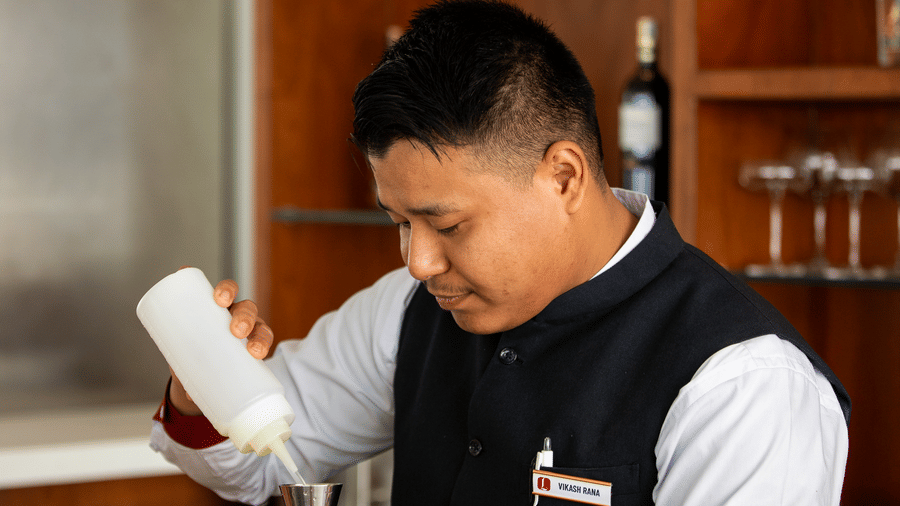 A bartender in uniform prepares a drink behind the counter, pouring liquid into a jigger, with bar tools and glasses neatly arranged at Mandala Norling Lords Prime in Kathmandu.