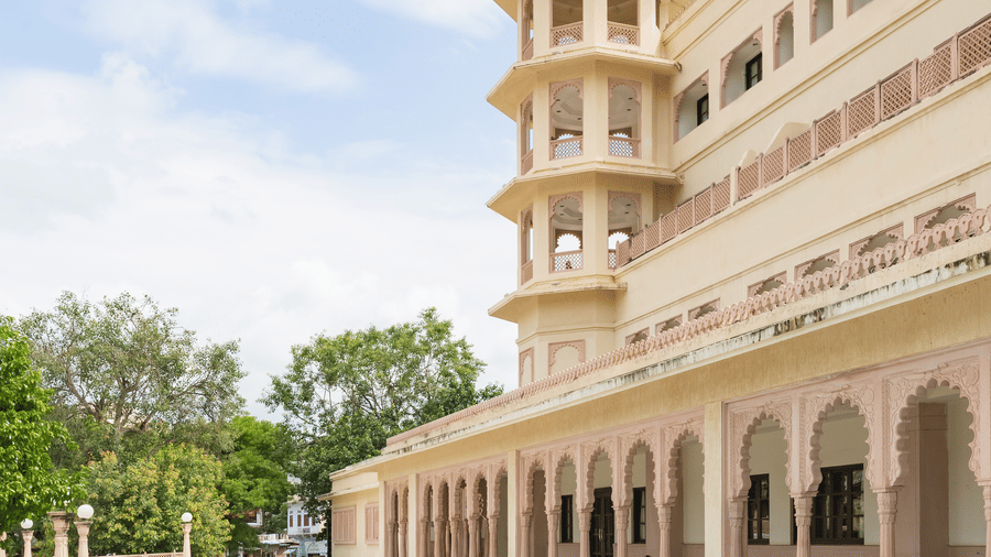 The facade of Shri Vallabh Vislas Lords Plaza Nathdwara featuring patterned white tiles in the foreground and lush green vegetation under blue sky at the backdrop.