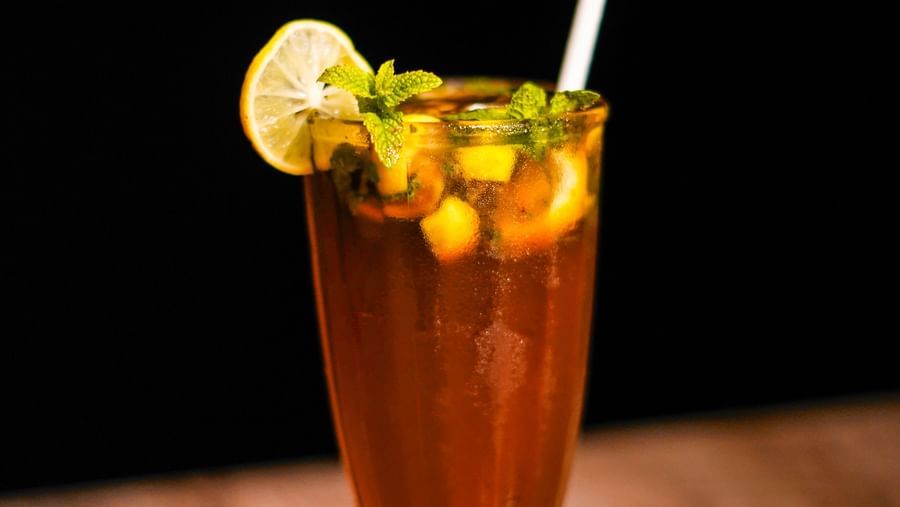 A tall glass filled with a dark beverage, garnished with a lime slice and a straw, on a wooden surface with a dark background | Sukama Resort & Spa, Nainital