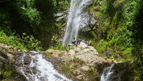 An image of a waterfall cascading through the rocks amidst the lush greenery of the forest with people enjoying the clean stream of water