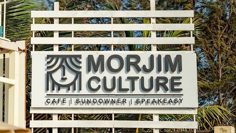 A white slatted sign at Living Room Hotels featuring MORJIM CULTURE in black text and a stylised eye logo, set against a bright blue sky and green foliage. Below, smaller text reads CAFE . GALLERIE . APARTMENTS.