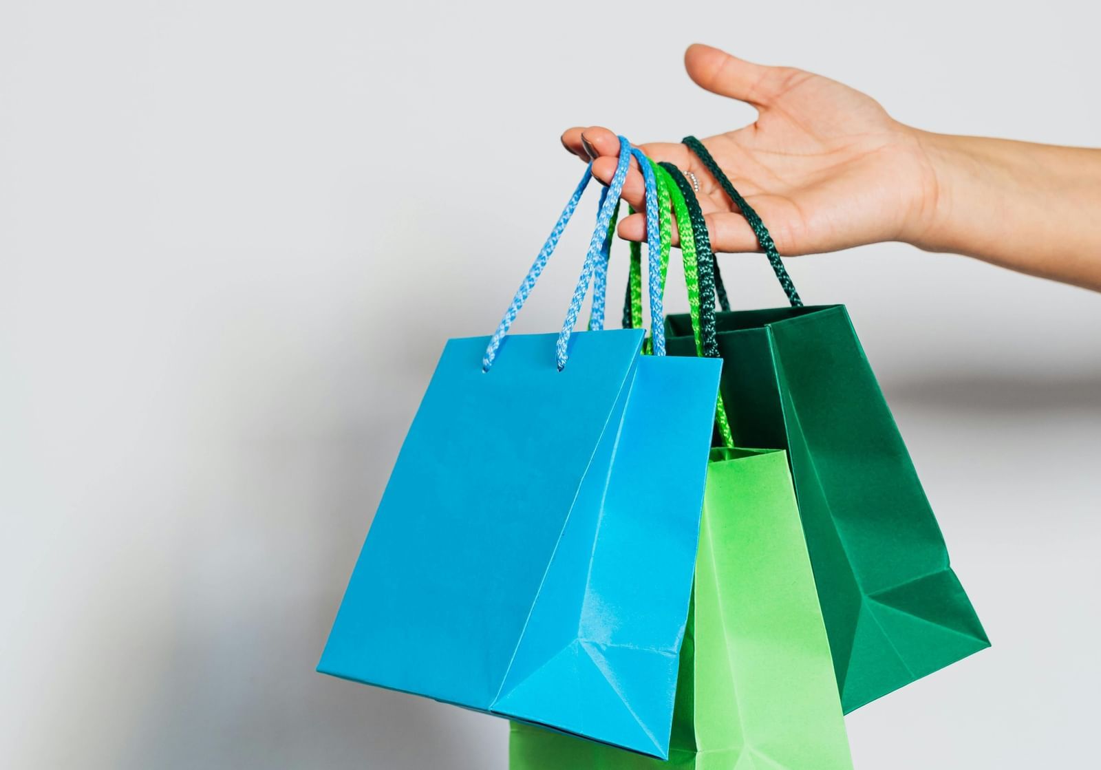 A person's hand stretched out, holding three paper shopping bags with a plain wall in the background.
