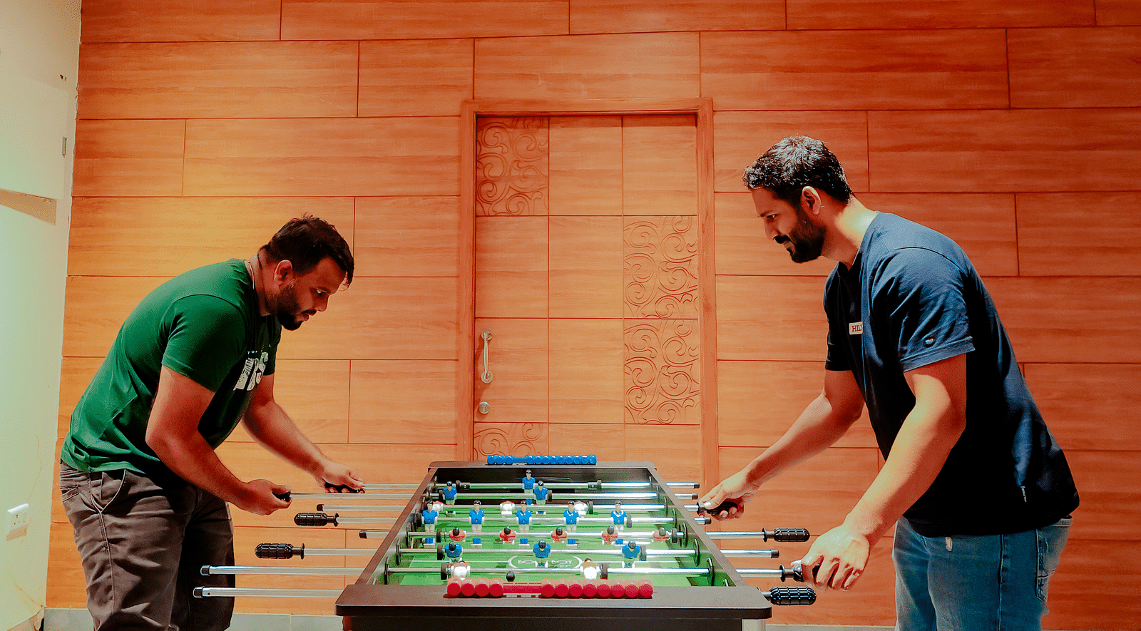 Two people play foosball in a room with brick walls and a high ceiling - Nirantara Resort & Spa.