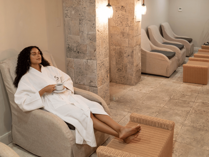 A person in a white robe relaxes on a cushioned chair, holding a mug, in a serene spa lounge with soft lighting, wicker footrests, and warm beige tones.