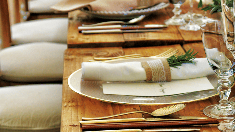A long wooden table with beautifully set cutlery with small fern leaves in vases.