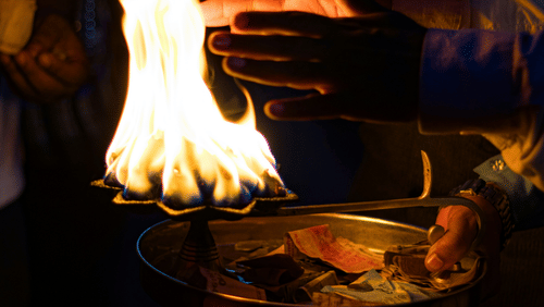 A person holding a metal lamp with a burning flame during a ritual inside a dimly lit temple area.
