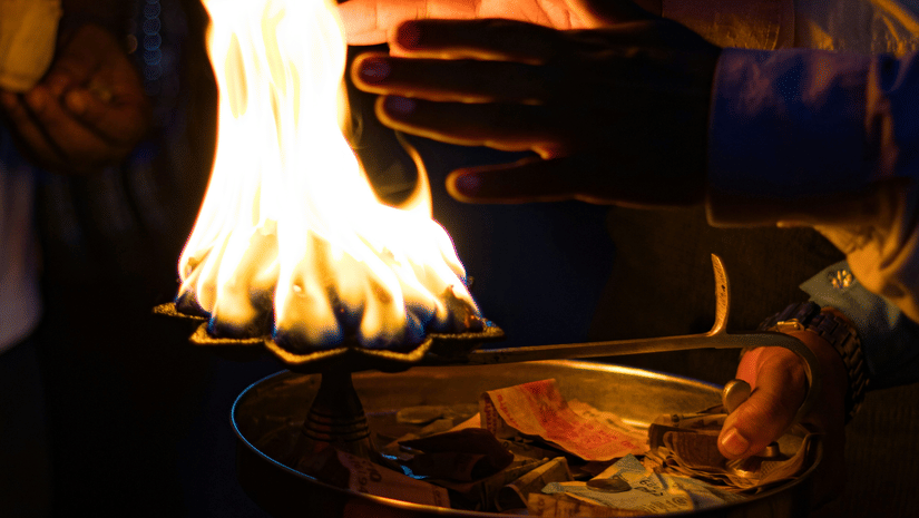 A person holding a metal lamp with a burning flame during a ritual inside a dimly lit temple area.