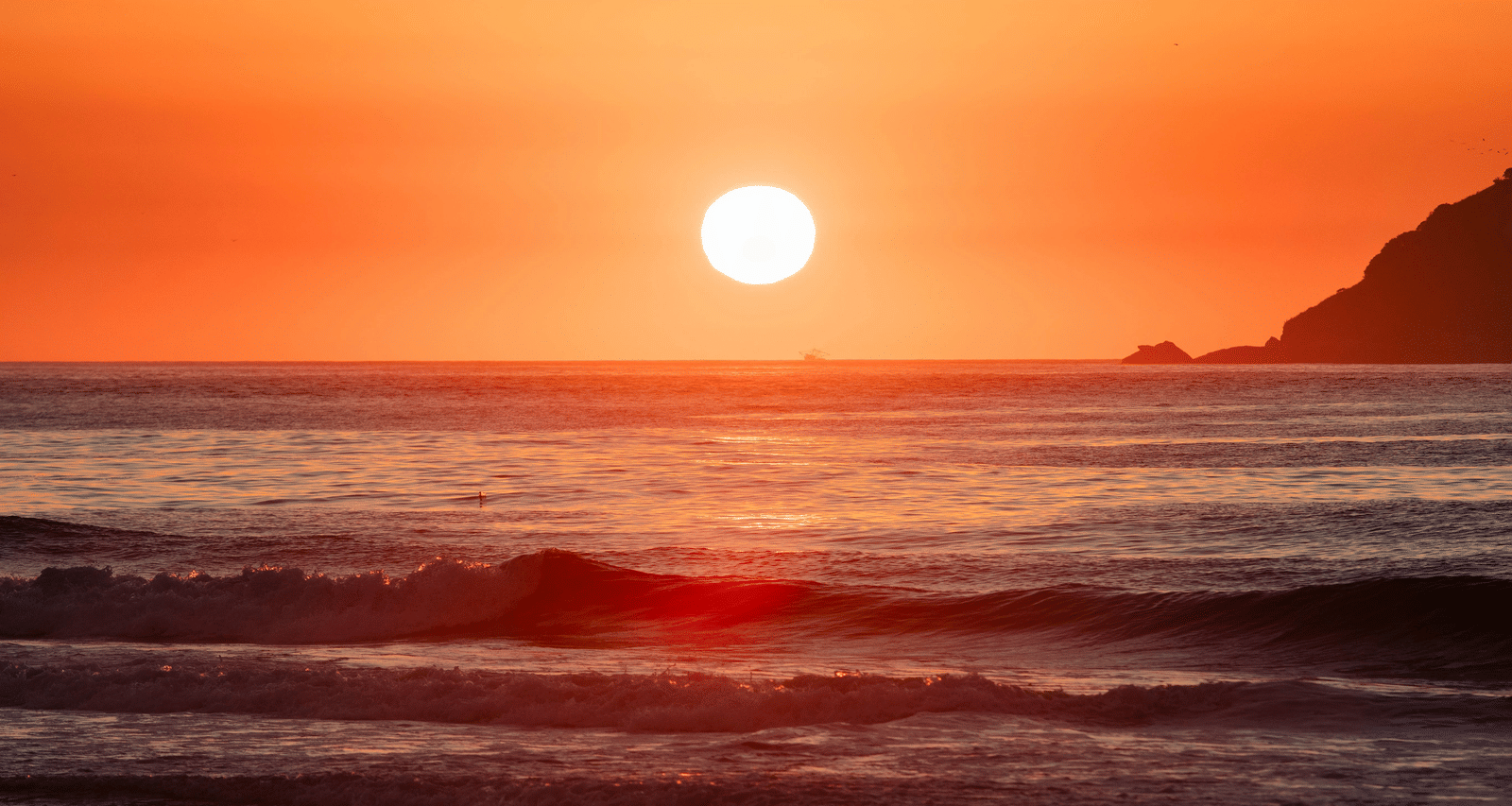 A bright orange sunset visible over the ocean, reflecting light onto the wet sand of a beach.