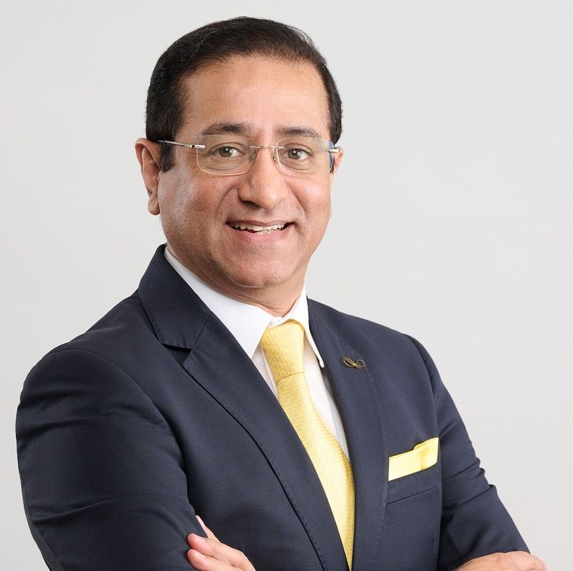 Nipun Vig in a black suit and white shirt with yellow tie, standing with arms crossed, smiling slightly against a plain background.