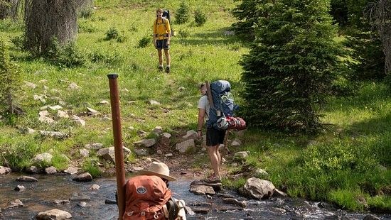 A trio climbing up a hill with backpacks, while passing a small stream of water.