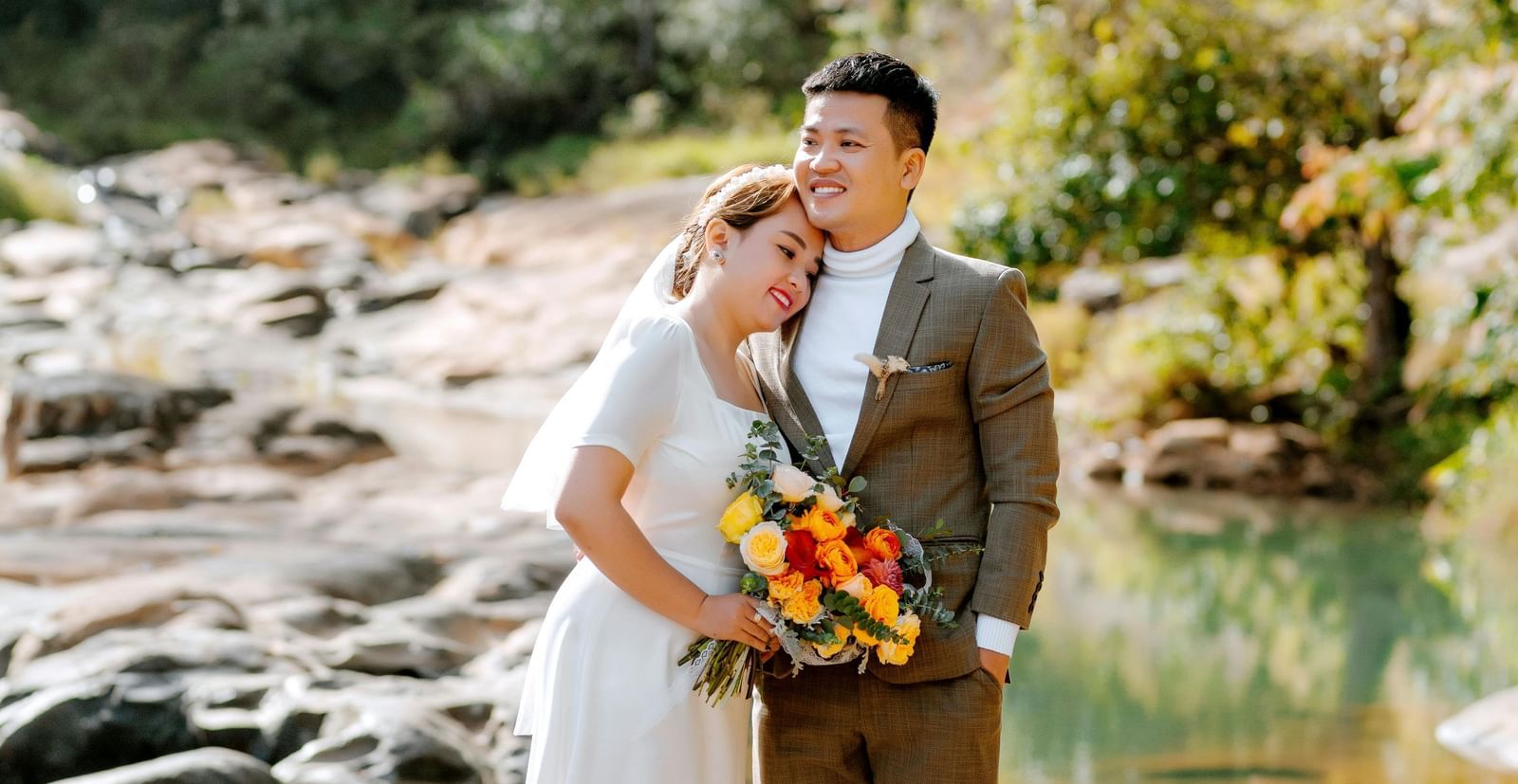 A bride and groom stand by a flowing river in a natural, outdoor setting, looking at each other