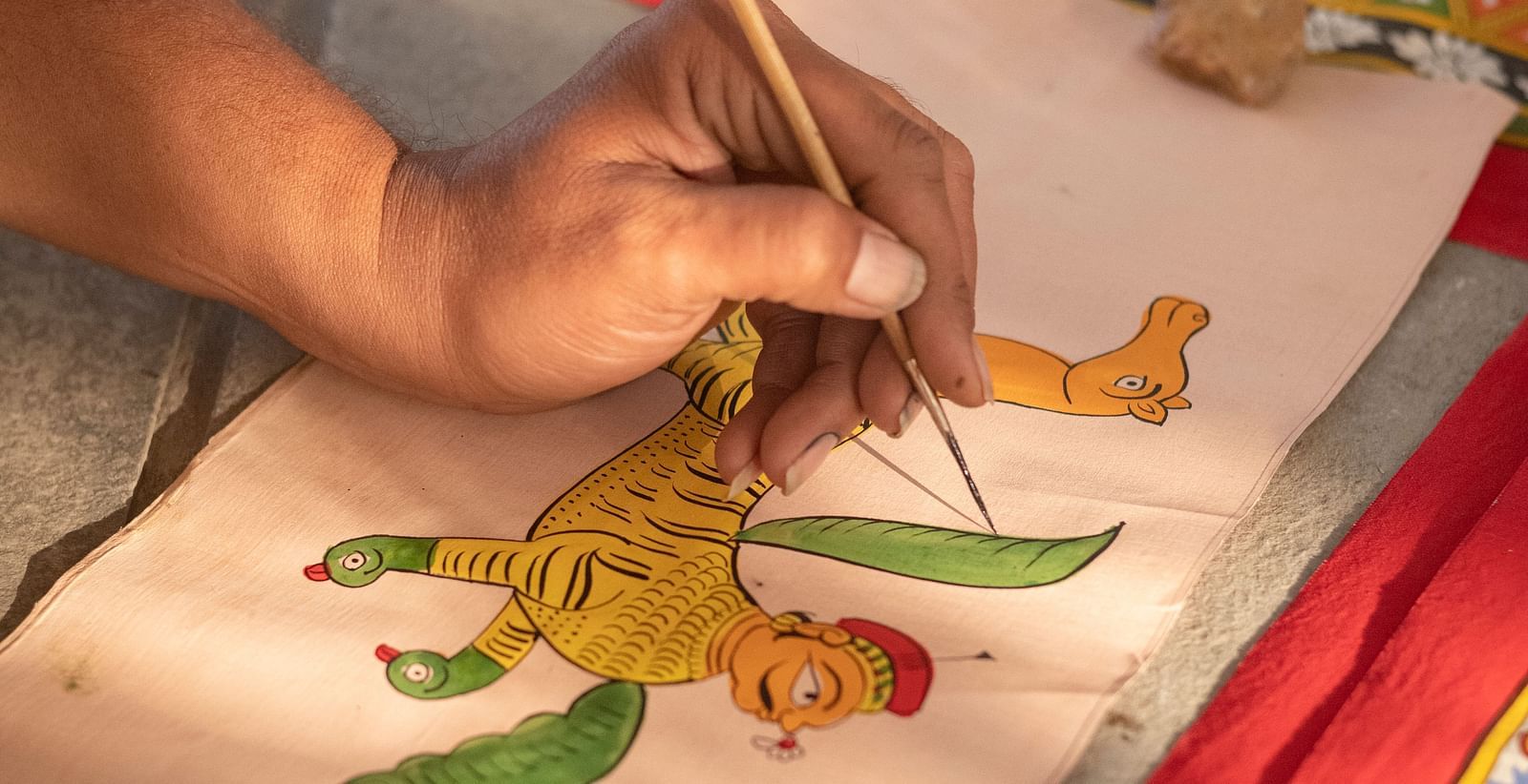 A person's hand painting a figure on a piece of cloth with a brush, with another decorated cloth visible in the background- Shahpura Bagh.