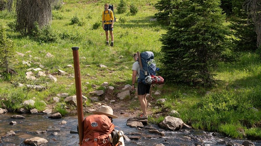 A trio climbing up a hill with backpacks, while passing a small stream of water.
