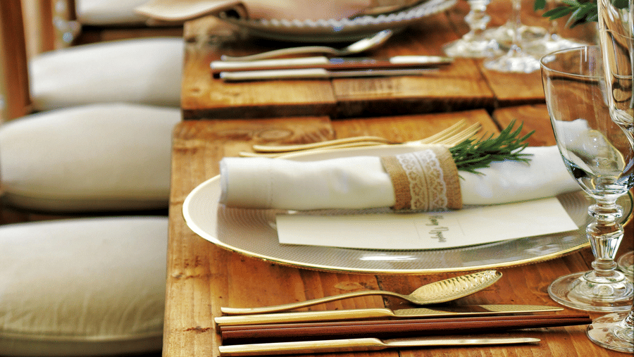 A long wooden table with beautifully set cutlery with small fern leaves in vases.