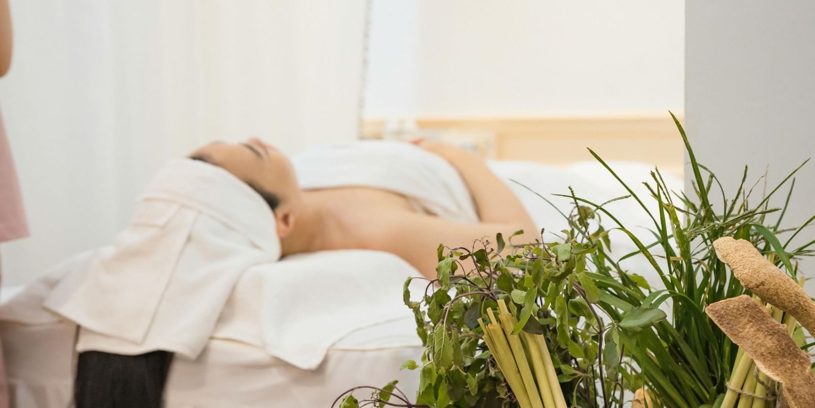 Woman relaxing on a spa bed with white towels, beside a basket of fresh herbs and a lit candle creating a calming ambiance.