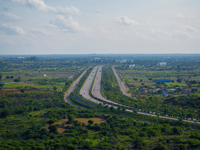 An aerial view of a wide, multi-lane highway stretching through a lush, green landscape under a bright, partly cloudy sky.