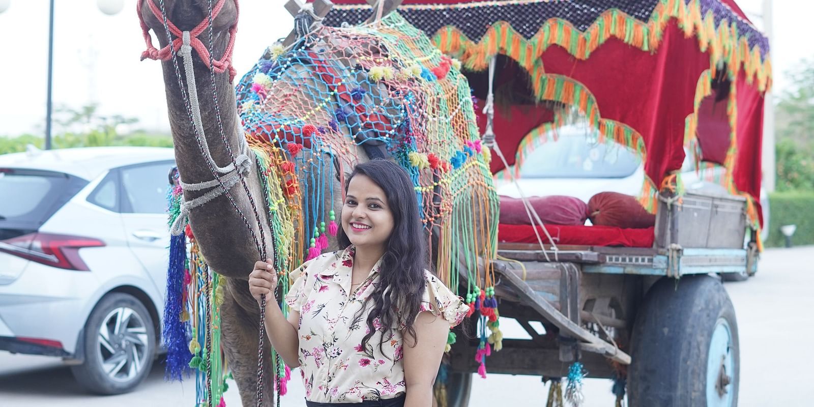 A woman stands next to a decorated camel and a small, two-wheeled carriage with a red canopy - Bamboo Saa Mulberry Resort, Pushkar
