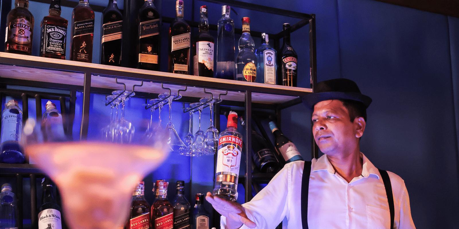 A bartender standing behind a bar with rows of bottles, illuminated by strong blue and purple nightclub lighting at Hotel Hukam's Lalit Mahal.