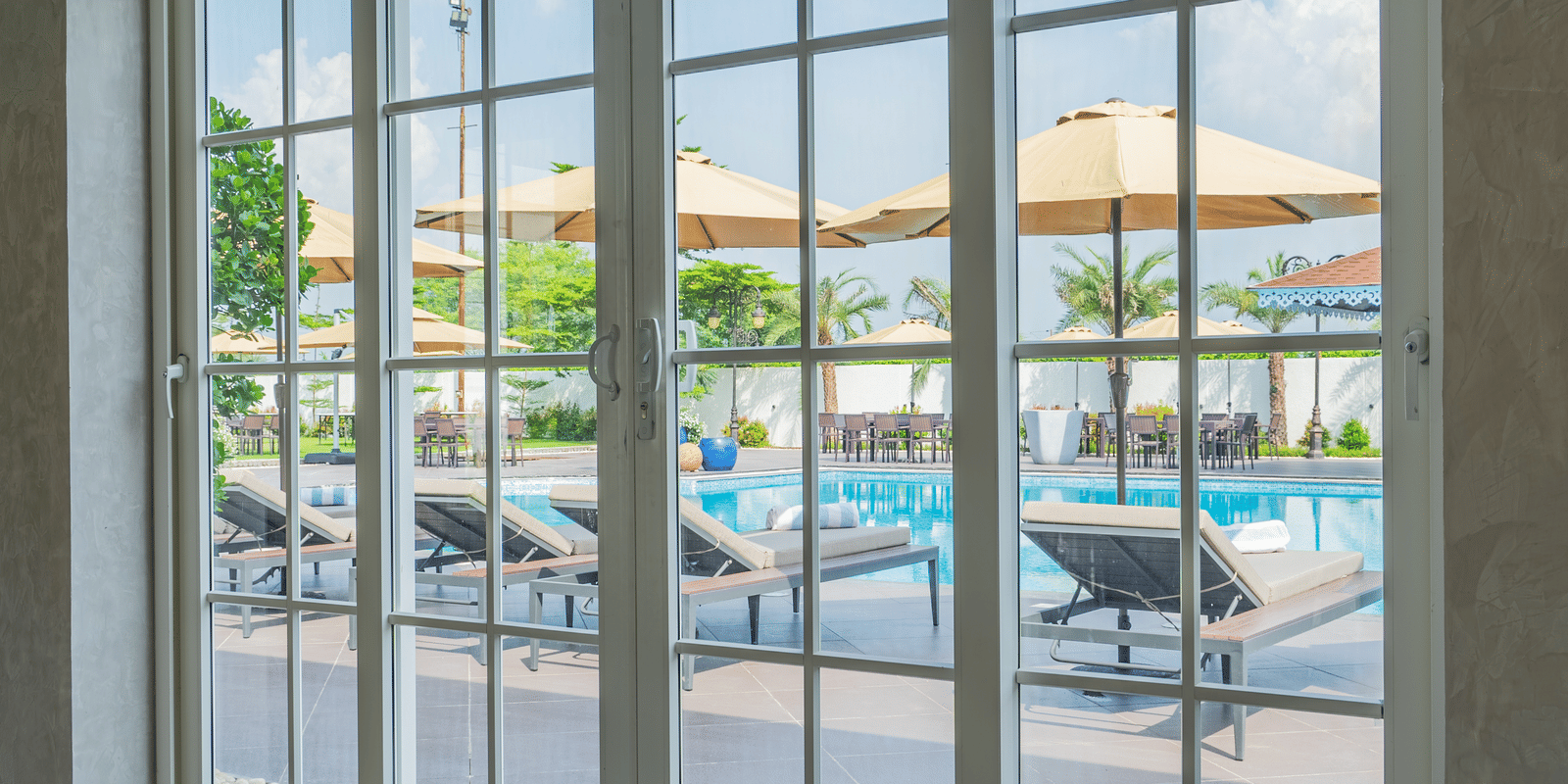 Interior view through large, gridded windows, showing a tiled floor and the outdoor pool area with deck chairs at Hotel Hukam's Lalit Mahal.