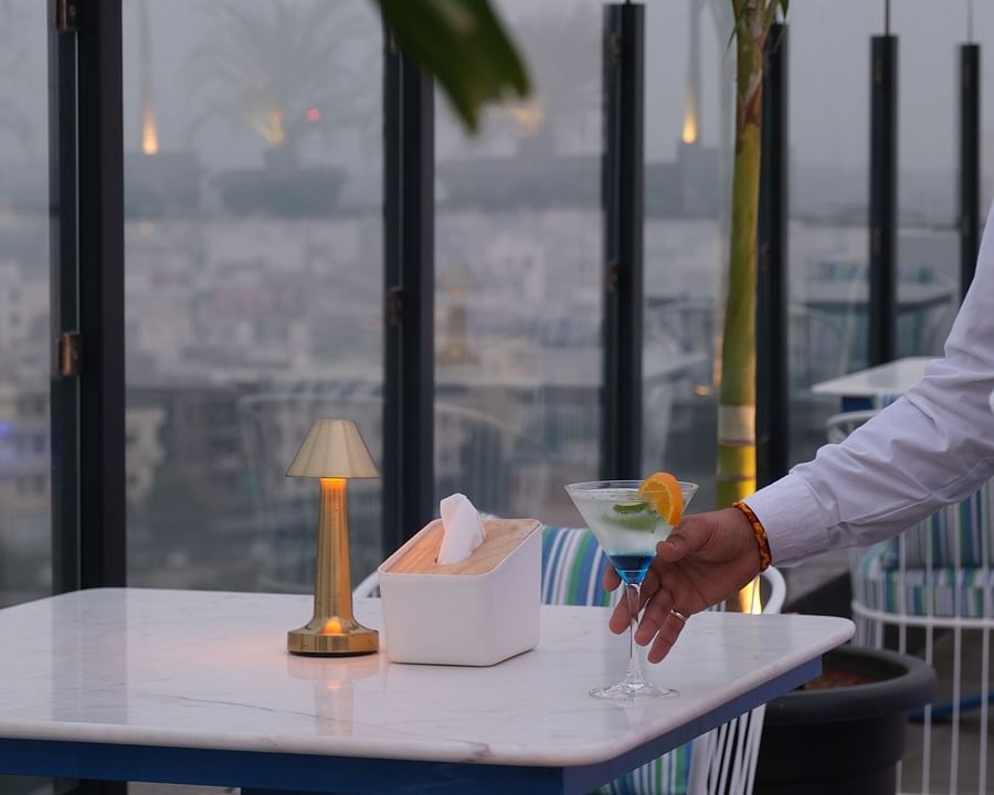 A Portrait of a server placing a Cocktail on a white table at ta BLU Rooftop Bar at Clarks Amer, Jaipur