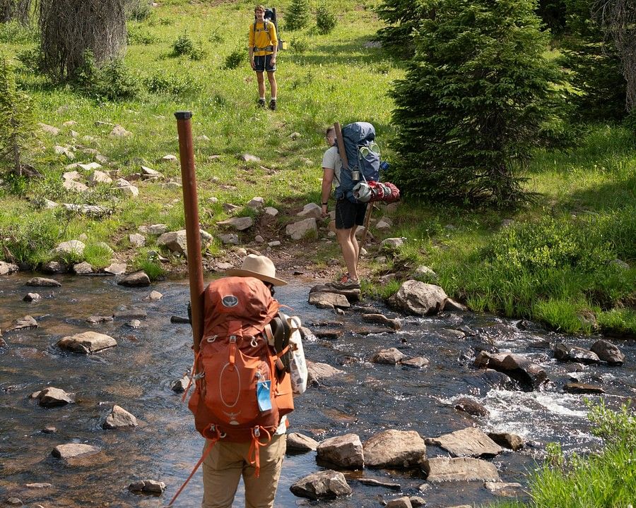 A trio climbing up a hill with backpacks, while passing a small stream of water.