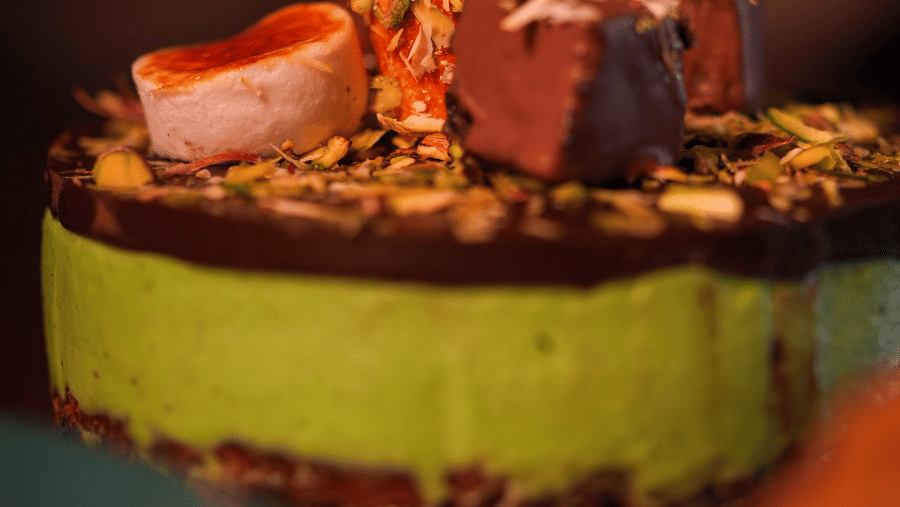 A vibrant dessert features a layered green and chocolate cake, topped with chocolate cubes, nuts, and colorful sprinkles, evoking a sense of indulgence.