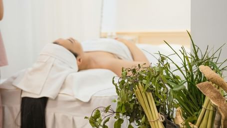 Woman relaxing on a spa bed with white towels, beside a basket of fresh herbs and a lit candle creating a calming ambiance.
