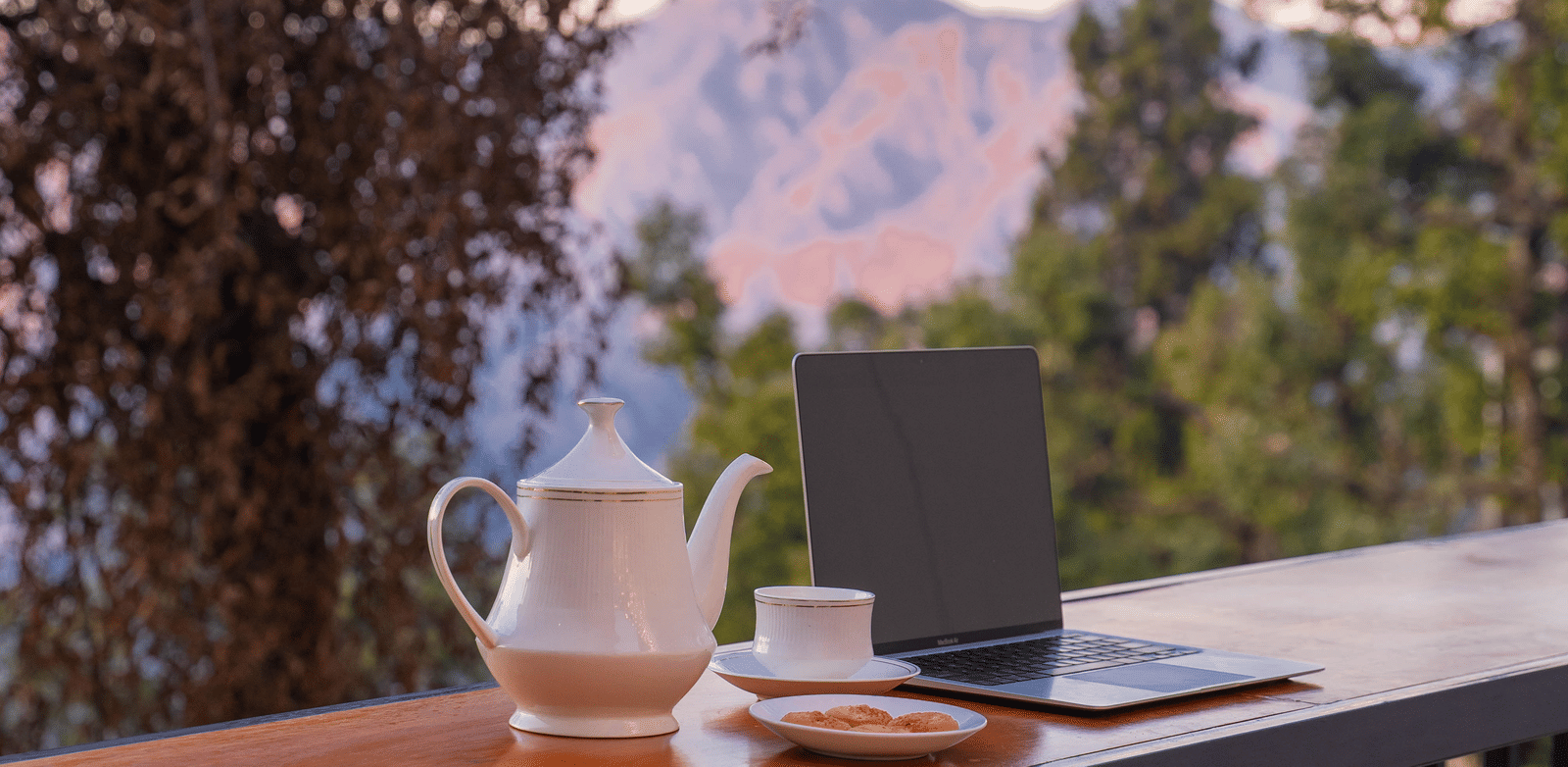 Close-up shot of a laptop along with a tea pot and a cup at Perfectstayz Premium Westend Mall Road, Mussoorie, with a beautiful backdrop of the mountains.