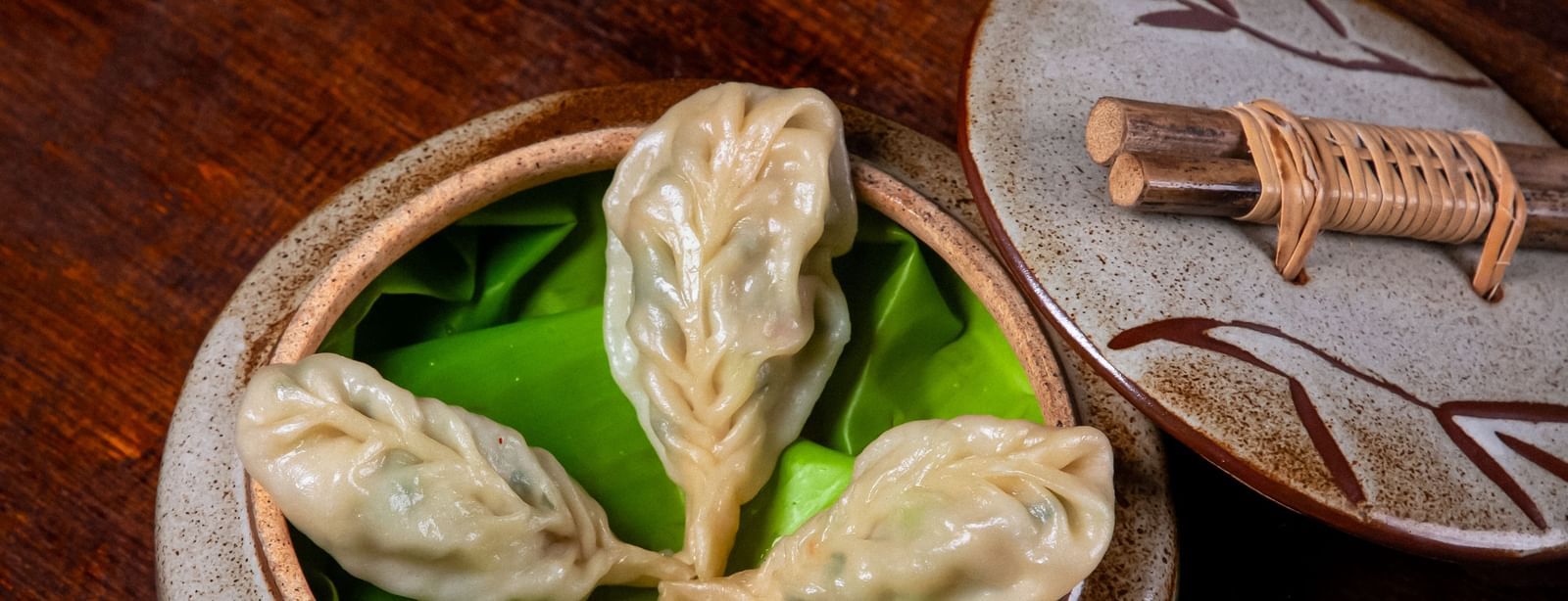  A bamboo steamer containing dumplings, garnished with herbs, accompanied by a spoonful of dipping sauce on a dark wooden table with lemongrass and green onions at The Hamuse Hotel, Kodaikanal.